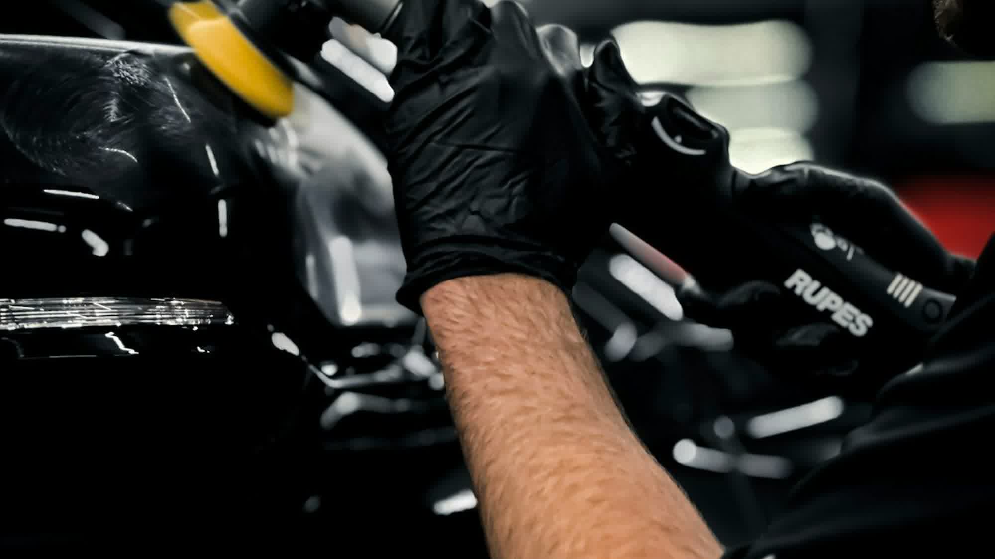 A gloved hand working a dual-action polisher across a dark panel under bay lighting