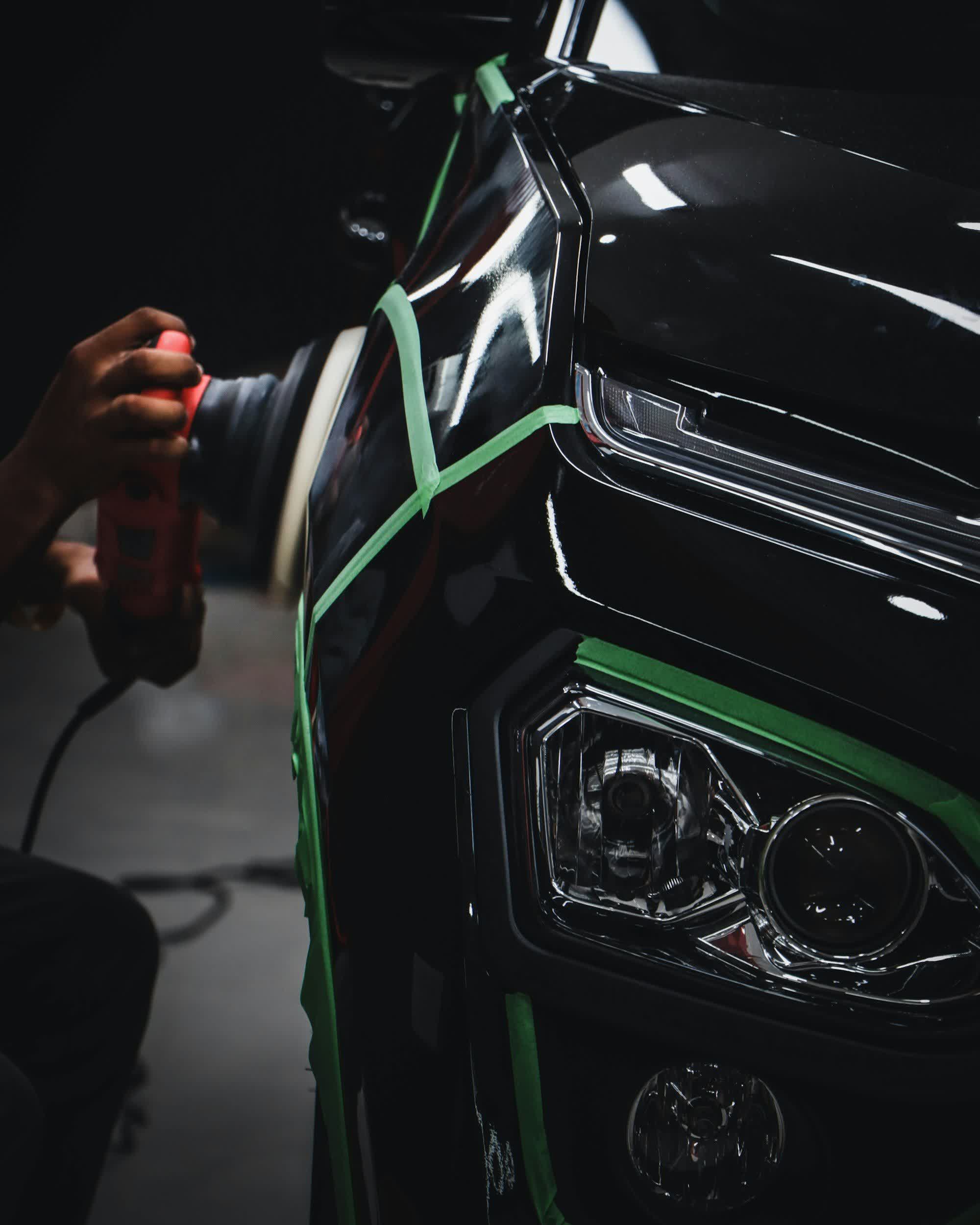 Masked panel edges beside a headlight with a polisher held ready in a dark bay